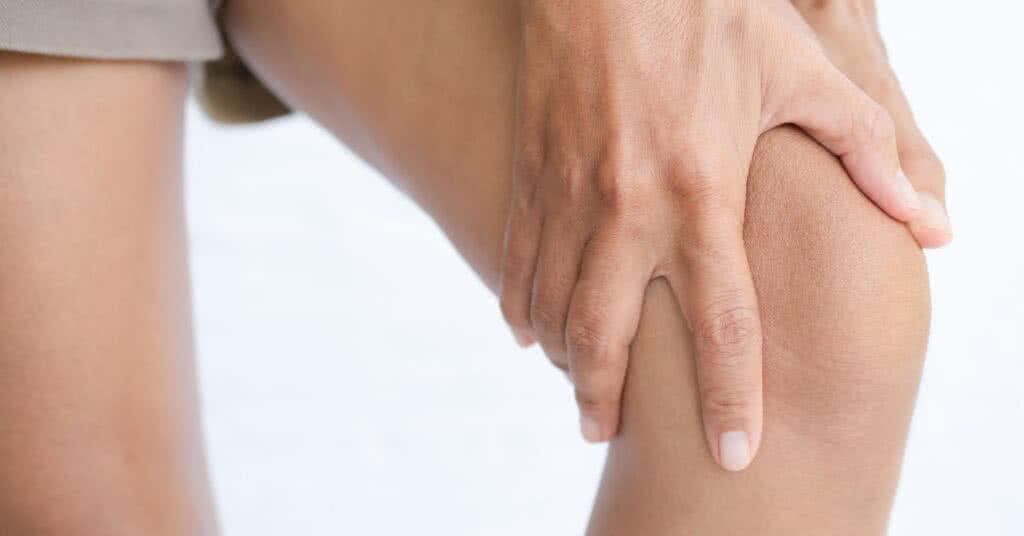 Imagem de uma mulher segurando o joelho com expressão de dor. A dor nas pernas pode ser um sinal de problemas vasculares, como varizes ou trombose. É importante procurar um médico para avaliar a causa da dor e iniciar o tratamento adequado.