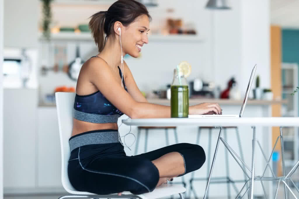 Mulher jovem e esportiva trabalhando em seu laptop após sessão de exercícios na cozinha de casa.