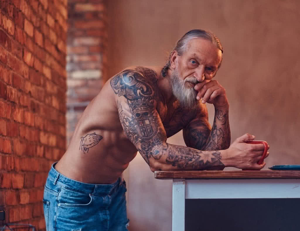 Imagem de um homem idoso musculoso com tatuagens no corpo, em um ambiente interno. Ele está em pé, exibindo seus músculos e tatuagens. Esta imagem é relevante para o post "Tatuagem masculina e saúde", que aborda os cuidados necessários para manter a saúde ao fazer tatuagens.